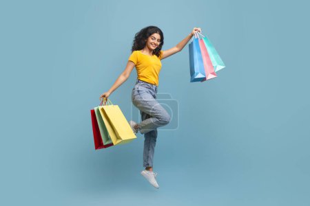 Happy hindu woman jumping high with shopping bags in her hands, cheerful positive young indian lady having fun against blue background, enjoying seasonal sales and discounts, copy space