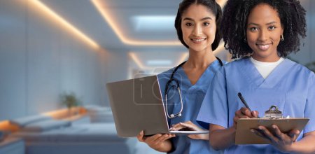 Two women nurses, one holding a laptop and the other writing on a clipboard, are smiling confidently in a modern hospital setting, symbolizing technological advancement in healthcare