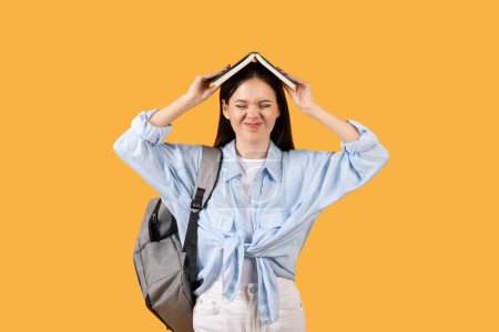 Foto de Estudiante cansada con libro en la cabeza y mueca, llevando mochila, de pie sobre fondo de estudio amarillo - Imagen libre de derechos