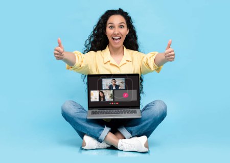 Foto de Una joven alegre con una camisa amarilla da un doble pulgar hacia arriba mientras está sentada con las piernas cruzadas con una computadora portátil que muestra una videollamada con participantes sonrientes sobre un fondo azul - Imagen libre de derechos