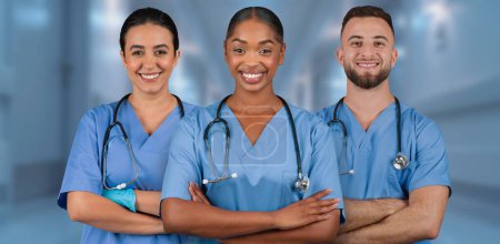 Téléchargez les photos : Trois professionnels de santé souriants internationaux en blouse bleue avec stéthoscopes se tiennent en confiance avec les bras croisés dans un couloir hospitalier bien éclairé, dépeignant le travail d'équipe - en image libre de droit