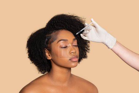 Photo for Peaceful African American woman with eyes closed receiving facial serum treatment applied with dropper by cosmetologist wearing gloves, set against soft beige background - Royalty Free Image