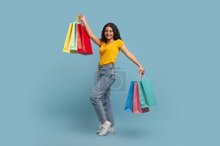 Seasonal Sales And Shopping. Full length of overjoyed young indian woman consumer holding many colorful bags with new purchases isolated on blue studio wall, copy space for advert