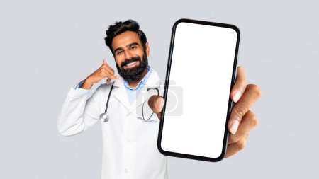 Téléchargez les photos : Joyeux médecin indien barbu en manteau blanc gestes appelez-moi signe tout en présentant smartphone avec écran d'espace de copie, isolé sur fond gris, annonce médicale - en image libre de droit