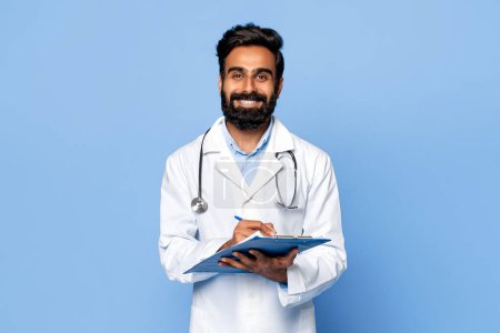 Téléchargez les photos : Médecin indien barbu souriant écrivant sur presse-papiers, équipé d'un stéthoscope, vêtu d'une blouse blanche sur fond bleu vif - en image libre de droit
