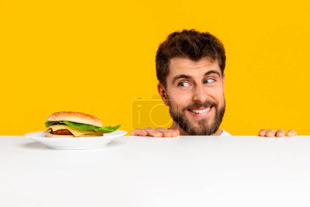 Photo for Young bearded guy looking at delicious burger on plate in studio with yellow background, struggling between guilty pleasure and desire for balanced healthy nutrition. Junk food temptation - Royalty Free Image