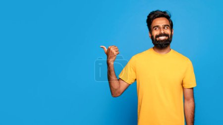 Photo for Happy bearded indian man in yellow shirt smiling and pointing with thumbs aside at free space, standing against vibrant blue background, panorama - Royalty Free Image