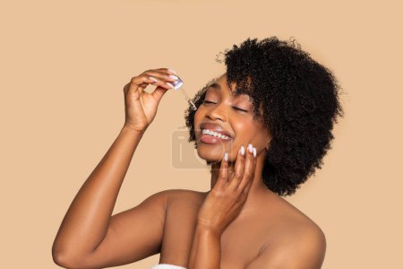 Foto de Joven mujer afroamericana alegre con los ojos cerrados, aplicando aceite facial con gotero, sonriendo y tocándose la cara suavemente, disfrutando de su rutina de belleza sobre fondo beige - Imagen libre de derechos