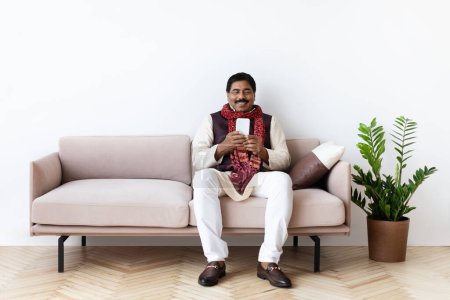Téléchargez les photos : Heureux homme indien d'âge moyen portant des vêtements traditionnels assis sur le canapé dans le salon confortable intérieur, en utilisant un téléphone portable à la maison, lire les nouvelles en ligne, défilement, websurf, espace de copie - en image libre de droit