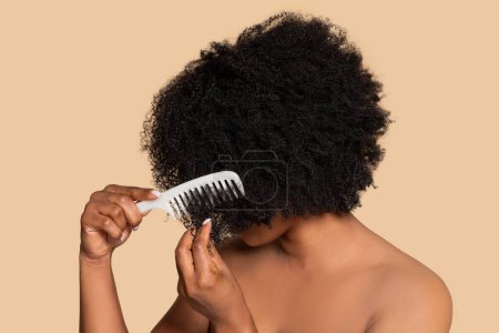 Foto de Mujer afroamericana joven desenreda su cabello rizado rico con peine, posando sobre un telón de fondo neutro, simbolizando el cuidado diario de la belleza - Imagen libre de derechos