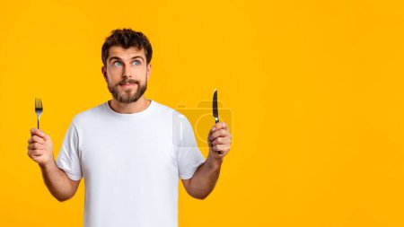 Photo for Hungry bearded European guy holds knife and fork ready to savor restaurant meal, standing over yellow background in studio. Hunger and food cravings. Panorama with empty space - Royalty Free Image