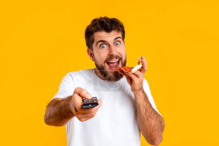 Photo for Excited millennial man switches channels on TV with remote control while biting pizza slice, studio portrait over yellow background. Pleasure of fast food, binge watching and eating - Royalty Free Image