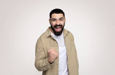 Photo for Cheerful shocked handsome young latin man with beard in casual with open mouth, scream, rise fist up, isolated on gray background, studio. Human emotions, surprise, win and reaction at good news - Royalty Free Image