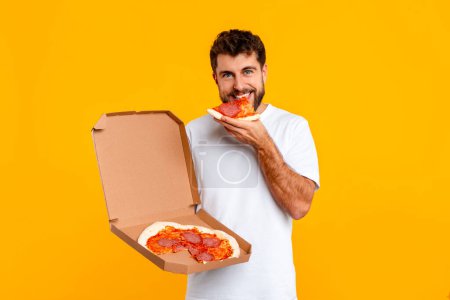 Photo for Hungry young European man bites into slice of pizza, emphasizing concept of comfort eating and junk food consumption, standing on yellow background, looking at camera. Nutrition habits - Royalty Free Image