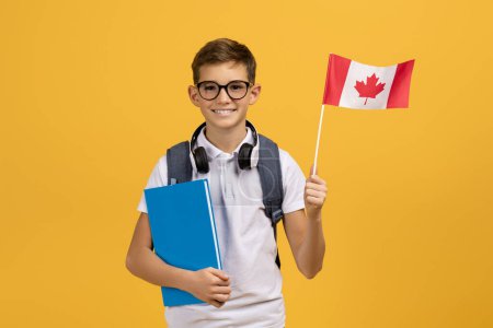 Photo for International Education. Happy Teen Boy With Backpack Holding Canadian Flag And Workbooks, Portrait Of Smiling Schoolboy Posing On Yellow Studio Background, Enjoying Study Abroad, Copy Space - Royalty Free Image