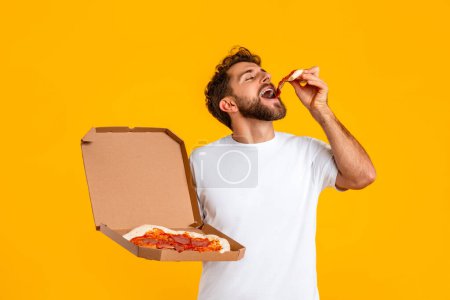 Foto de Millennial hombre en camiseta blanca satisface sus antojos con comida rápida, la celebración de la caja de entrega y disfrutar de rebanada de pizza en medio de fondo de estudio amarillo. Concepto hábitos alimenticios, comida de consuelo - Imagen libre de derechos
