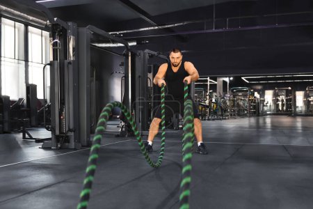 Photo for Crossfit training. Motivated muscular man exercising with battle ropes at gym, young caucasian male athlete engaging in high-intensity workout, challenging his strength and endurance in sport club - Royalty Free Image