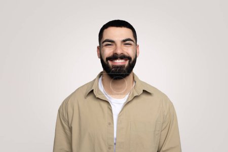 Foto de Sonriente joven latino guapo con barba en casual mirando a la cámara, aislado sobre fondo gris, estudio. Emociones humanas, retrato, fotos y videollamadas, estilo de vida positivo - Imagen libre de derechos