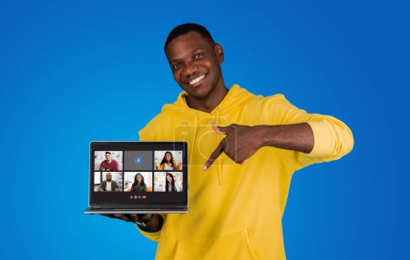Téléchargez les photos : Un homme souriant dans un sweat à capuche jaune pointe vers un écran d'ordinateur portable affichant une vidéoconférence avec divers participants sur un fond bleu audacieux. Recommandation de réunion, app - en image libre de droit
