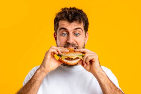 Photo for Caucasian man happily biting into a burger, portrait shot over yellow studio background, indulging in junk food, posing with delicious cheeseburger. Fast food enjoyment, nutrition habits - Royalty Free Image