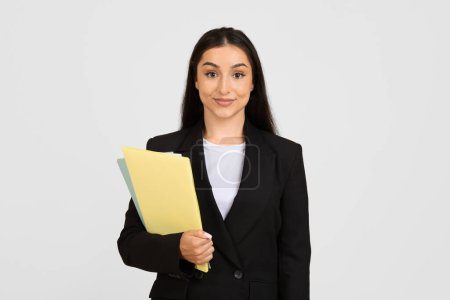 Photo for Professional young businesswoman in black suit, confidently holding colorful folders, ready for meeting, posing against clean grey background - Royalty Free Image