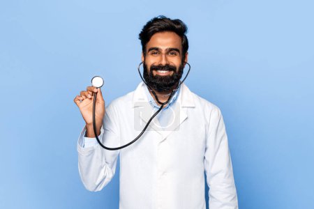 Foto de Radiante médico indio de mediana edad con bata blanca de laboratorio usando estetoscopio, con expresión alegre, posando sobre fondo azul brillante - Imagen libre de derechos