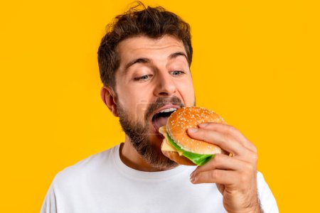 Téléchargez les photos : Homme appréciant cheeseburger le jour de la triche, ayant des envies de restauration rapide sur fond jaune vif. Portrait rapproché de jeune homme barbu prenant morsure de hamburger savoureux en studio. Réconfort alimentaire - en image libre de droit