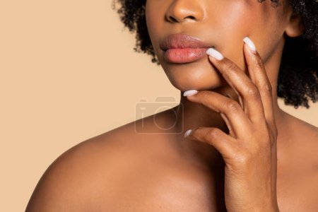 Foto de Primer plano de la mujer contemplativa afroamericana tocándose suavemente los labios, con una sonrisa sutil, demostrando su belleza natural contra un fondo beige neutro - Imagen libre de derechos