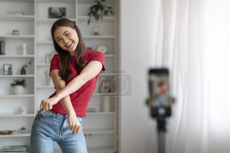 Foto de Concepto de Blogging. Mujer asiática alegre bailando en la cámara, joven coreana haciendo danza divertida mientras graba video en casa, mujer positiva usando teléfono inteligente en el trípode, espacio libre - Imagen libre de derechos