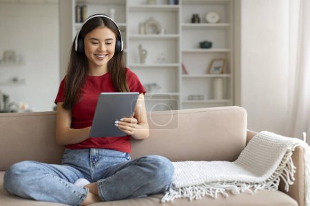 Foto de Mujer asiática sonriente usando auriculares usando tableta digital en casa, mujer coreana feliz sentada con las piernas cruzadas en el sofá en la sala de estar bien iluminada, retratando la relajación y la interacción digital - Imagen libre de derechos