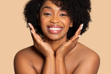 Téléchargez les photos : Femme afro-américaine souriante rayonnante avec les mains encadrant délicatement son visage, mettant en valeur la beauté naturelle et le bonheur sur fond beige - en image libre de droit