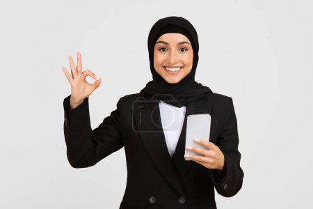 Photo for Cheerful Muslim businesswoman in hijab holding smartphone in one hand and making okay gesture with the other, symbolizing approval or success, against light background - Royalty Free Image