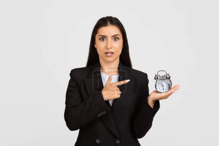 Photo for Concerned young businesswoman in black suit pointing at alarm clock shes holding, illustrating time management, deadlines, and urgency on light grey background - Royalty Free Image