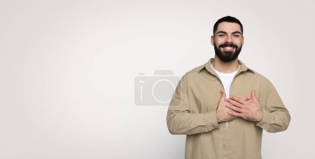 Téléchargez les photos : Heureux confiant beau jeune homme du Moyen-Orient avec une barbe décontractée mettre les mains à la poitrine du cœur, isolé sur fond gris, studio, panorama. Merci signe et geste de gratitude - en image libre de droit
