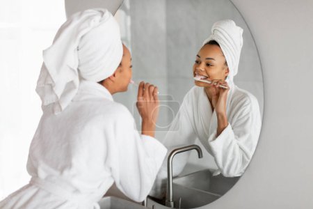 Téléchargez les photos : Soins dentaires. Femme noire joyeuse se brosse les dents et sourit au reflet dans le miroir, portant un peignoir de spa avec turban serviette, maintenant l'hygiène buccodentaire dans un cadre de salle de bain moderne - en image libre de droit