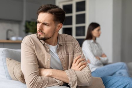 Photo for Distressed young man with arms crossed looking away from woman sitting on background out of focus after quarrel, implying strained relationship - Royalty Free Image