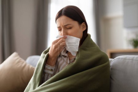 Unwell young woman covered with green blanket, using tissue to blow her nose, possibly suffering from cold or flu while sitting on couch at home