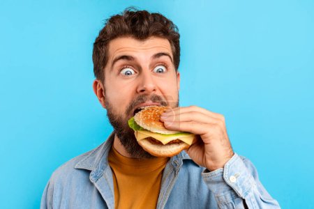 Téléchargez les photos : Homme caucasien affamé sur fond bleu mordant excitamment dans un hamburger, révélant le plaisir de se livrer à la malbouffe. Coup de tête d'un gars excité mangeant un cheeseburger. Fast food fringales concept - en image libre de droit