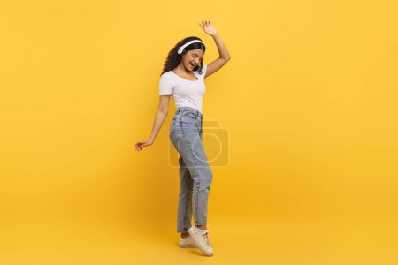 Foto de Joven estudiante india muy alegre usando ropa casual usando auriculares inalámbricos y bailando con los ojos cerrados, aislado en el fondo amarillo del estudio, espacio para copiar, longitud completa - Imagen libre de derechos