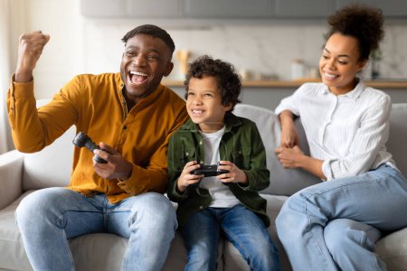 Téléchargez les photos : Joyeuse famille noire jouer à des jeux vidéo, avec le père célébrant la victoire, l'enfant engagé et la mère les acclamant, assis ensemble sur le canapé - en image libre de droit