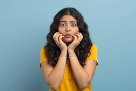 Photo for Appalled shocked pretty long-haired young indian woman wearing yellow t-shirt touching her face and grimacing, expressing strong emotions, isolated on blue studio background - Royalty Free Image