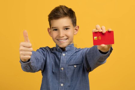 Photo for Cheerful teenage boy presenting red credit card and showing thumb up gesture, happy male child suggesting good banking services or approval, standing against bright yellow studio background - Royalty Free Image
