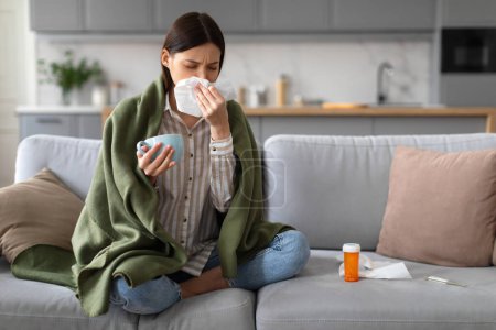 Foto de Mujer joven enferma sentada envuelta en manta, sonándose la nariz y sosteniendo la taza con bebida caliente, con medicamentos sobre la mesa, indicando resfriado o gripe en casa - Imagen libre de derechos