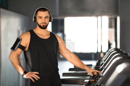Photo for Confident young man with headphones and armband phone holder standing by treadmills, handsome muscular male setting up for cardio session in modern gym, enjoying sport trainings, copy space - Royalty Free Image