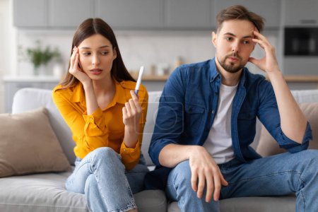 Photo for Young woman sits on couch, holding pregnancy test with contemplative look, while man next to her appears worried and pensive - Royalty Free Image