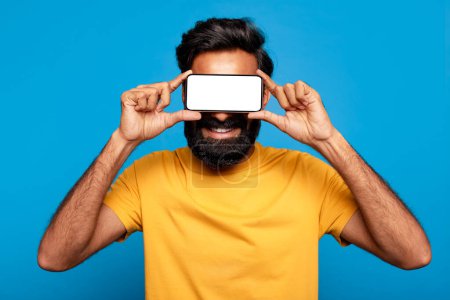 Photo for Playful indian bearded man in yellow t-shirt holding smartphone in front of his eyes like glasses, with beaming smile, on vivid blue background, mockup - Royalty Free Image
