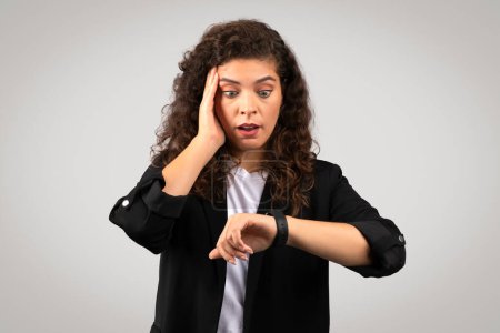 Photo for Shocked businesswoman checking time on her smartwatch and touching cheek, portraying sense of urgency, time management or deadline stress, gray background - Royalty Free Image