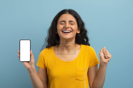 Photo for Great online offer. Emotional long-haired young indian lady wearing yellow t-shirt showing smartphone with white blank screen and gesturing with closed eyes, blue studio background, mockup, copy space - Royalty Free Image