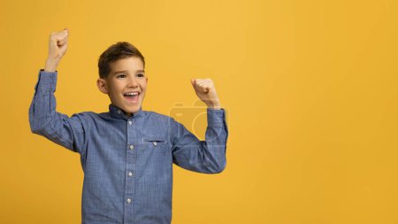 Foto de ¿Sí? Muchacho adolescente muy contento agitando puños celebrando el éxito mientras está parado sobre el fondo amarillo del estudio, niño masculino feliz emocional mirando al lado del espacio de copia con emoción, Panorama - Imagen libre de derechos