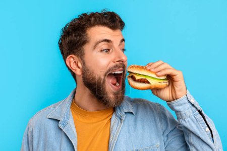 Photo for Headshot of young European man takes bite of tasty burger with joyful expression, enjoying delicious meal over blue studio backdrop. Concept of junk food and nutritional balance - Royalty Free Image